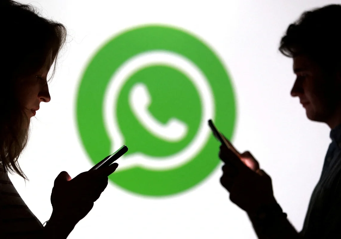 Um homem e uma mulher usam seus celulares simultaneamente. Há um logo do WhatsApp ao fundo, levemente desfocado. Falando sobre como fazer itálico no whatsapp.