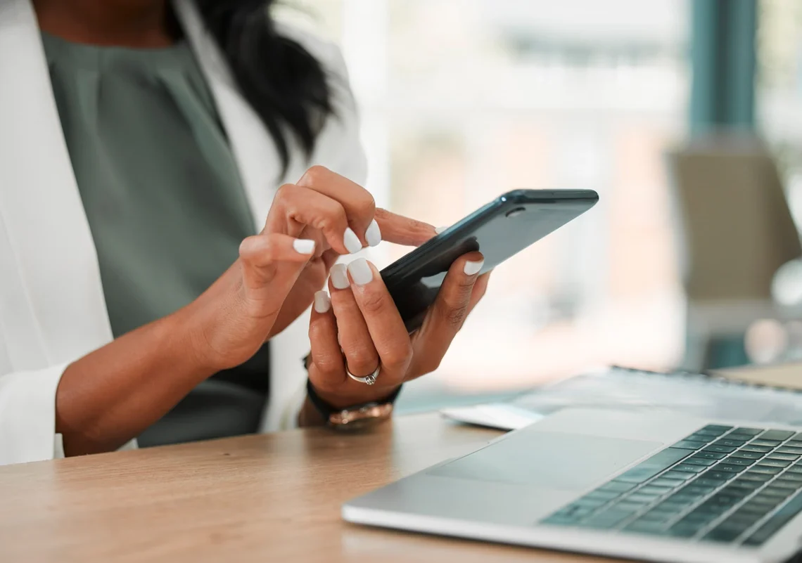 Mulher utilizando celular em frente a um notebook. Dando 5 dicas para ser mais produtivo com o uso de smartphones.