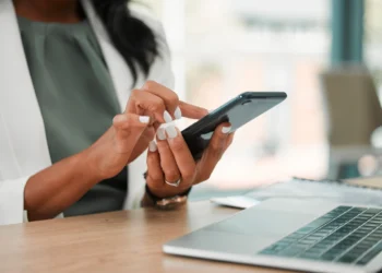 Mulher utilizando celular em frente a um notebook. Dando 5 dicas para ser mais produtivo com o uso de smartphones.